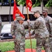 A U.S. Army change of command or activation ceremony is taking place outdoors, likely on a military installation in a tropical location. Two soldiers are exchanging a red unit guidon (flag marked 'HQ'), which is the central act of a formal military ceremony signifying transfer of command authority. Four soldiers in OCP (Operational Camouflage Pattern) uniforms are visible, with unit colors and a streamer-adorned organizational flag in the background.