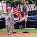 Two U.S. Army soldiers in MultiCam (OCP) uniforms stand at attention during a formal military ceremony, positioned in front of a color guard display. Multiple flags are visible including the U.S. national flag, a U.S. Army flag with streamers, and unit guidons. The setting appears to be an outdoor military installation, likely in a tropical location based on the palm trees and vegetation.