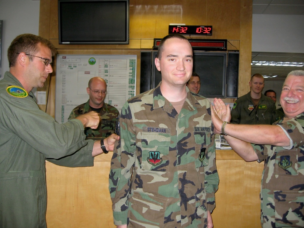 A U.S. Air Force military ceremony appears to be taking place in what looks like an operations center or command room. A service member named 'Steadman' (visible on name tape) is standing at attention while two other personnel appear to be conducting a coin or tapping ceremony on both sides. Several other military personnel are observing in the background.