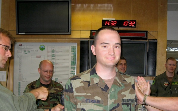 A U.S. Air Force military ceremony appears to be taking place in what looks like an operations center or command room. A service member named 'Steadman' (visible on name tape) is standing at attention while two other personnel appear to be conducting a coin or tapping ceremony on both sides. Several other military personnel are observing in the background.
