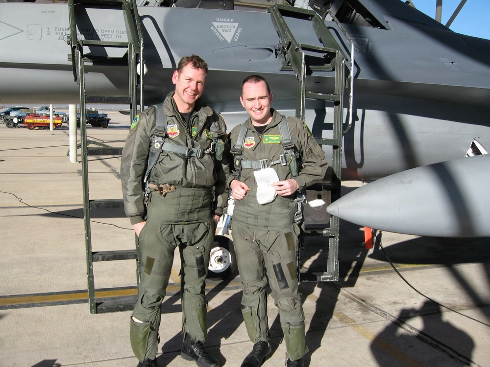 Two military aviators in olive green flight suits stand together on a flight line in front of a military jet aircraft. Both are wearing full flight gear including harnesses and patches on their suits, suggesting they have just completed or are preparing for a flight. The aircraft behind them displays a visible 'DANGER EJECTION SEAT' warning placard near the cockpit area.