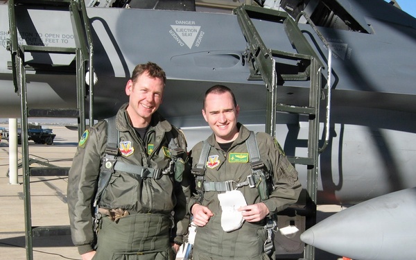 Two military aviators in olive green flight suits stand together on a flight line in front of a military jet aircraft. Both are wearing full flight gear including harnesses and patches on their suits, suggesting they have just completed or are preparing for a flight. The aircraft behind them displays a visible 'DANGER EJECTION SEAT' warning placard near the cockpit area.