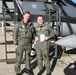 Two military aviators in olive green flight suits stand together on a flight line in front of a military jet aircraft. Both are wearing full flight gear including harnesses and patches on their suits, suggesting they have just completed or are preparing for a flight. The aircraft behind them displays a visible 'DANGER EJECTION SEAT' warning placard near the cockpit area.