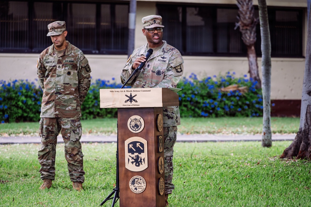 A U.S. Army soldier is speaking at a podium during an official military ceremony outdoors. The podium bears the insignia and text of the '94th Army Air &amp; Missile Defense Command' with the motto 'First Line of Defense.' A second soldier stands at ease in the background.