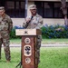 A U.S. Army soldier is speaking at a podium during an official military ceremony outdoors. The podium bears the insignia and text of the '94th Army Air &amp; Missile Defense Command' with the motto 'First Line of Defense.' A second soldier stands at ease in the background.