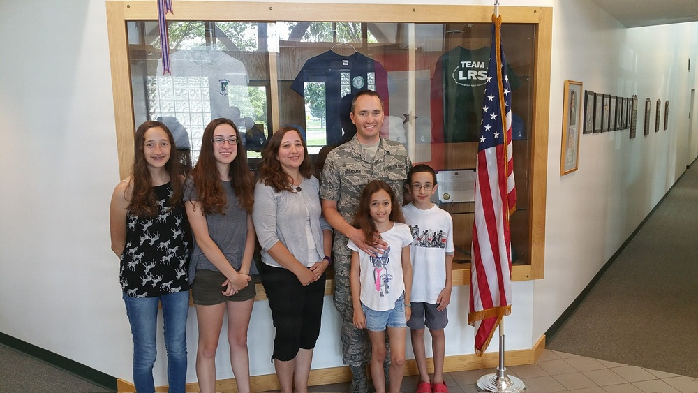 A group of six people, including a U.S. Air Force service member in camouflage uniform, pose together in what appears to be a military building hallway. They are standing in front of a display case containing military-themed shirts and memorabilia, with an American flag to their right. The group appears to be a family, consisting of two adults and four children of varying ages.