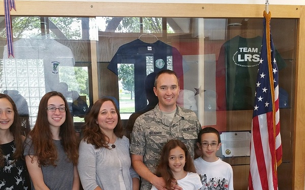 A group of six people, including a U.S. Air Force service member in camouflage uniform, pose together in what appears to be a military building hallway. They are standing in front of a display case containing military-themed shirts and memorabilia, with an American flag to their right. The group appears to be a family, consisting of two adults and four children of varying ages.