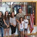 A group of six people, including a U.S. Air Force service member in camouflage uniform, pose together in what appears to be a military building hallway. They are standing in front of a display case containing military-themed shirts and memorabilia, with an American flag to their right. The group appears to be a family, consisting of two adults and four children of varying ages.