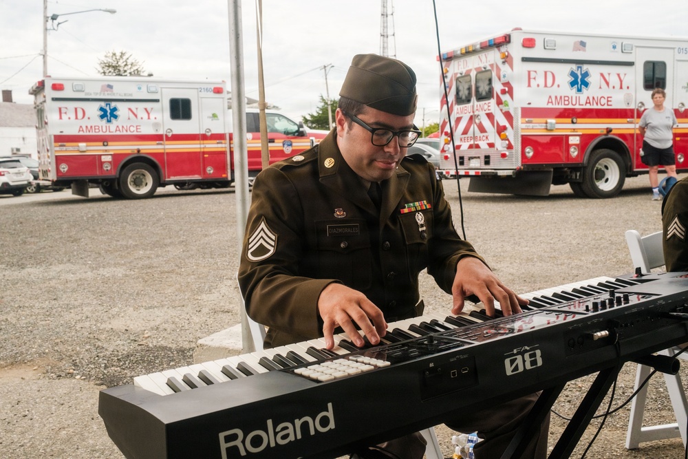 A U.S. Army staff sergeant in olive drab dress uniform is playing a Roland FA-08 keyboard synthesizer outdoors at what appears to be a public event. Multiple FDNY ambulances are visible in the background parking area. The soldier wears rank insignia, service ribbons, and a garrison cap.