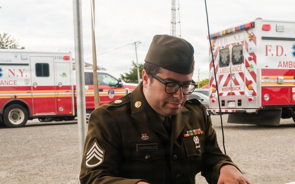 A U.S. Army staff sergeant in olive drab dress uniform is playing a Roland FA-08 keyboard synthesizer outdoors at what appears to be a public event. Multiple FDNY ambulances are visible in the background parking area. The soldier wears rank insignia, service ribbons, and a garrison cap.