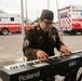 A U.S. Army staff sergeant in olive drab dress uniform is playing a Roland FA-08 keyboard synthesizer outdoors at what appears to be a public event. Multiple FDNY ambulances are visible in the background parking area. The soldier wears rank insignia, service ribbons, and a garrison cap.