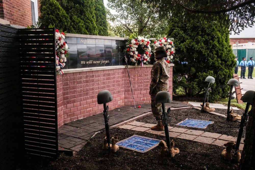 A military service member stands at attention before a September 11 memorial wall inscribed with 'We will never forget.' The memorial features battle cross displays (rifles, helmets, and boots) arranged in the foreground, flanked by red, white, and blue floral wreaths. Additional uniformed personnel are visible in the background, suggesting a formal commemorative ceremony.