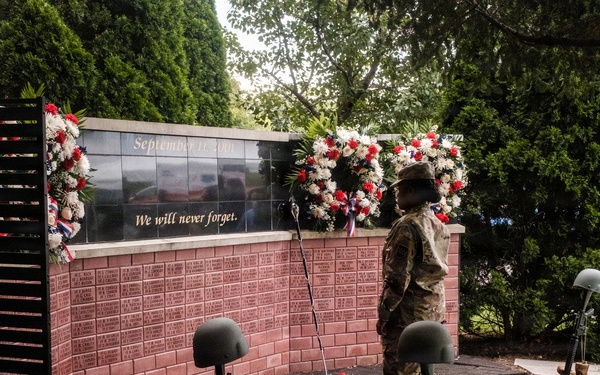 A military service member stands at attention before a September 11 memorial wall inscribed with 'We will never forget.' The memorial features battle cross displays (rifles, helmets, and boots) arranged in the foreground, flanked by red, white, and blue floral wreaths. Additional uniformed personnel are visible in the background, suggesting a formal commemorative ceremony.