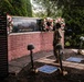A military service member stands at attention before a September 11 memorial wall inscribed with 'We will never forget.' The memorial features battle cross displays (rifles, helmets, and boots) arranged in the foreground, flanked by red, white, and blue floral wreaths. Additional uniformed personnel are visible in the background, suggesting a formal commemorative ceremony.