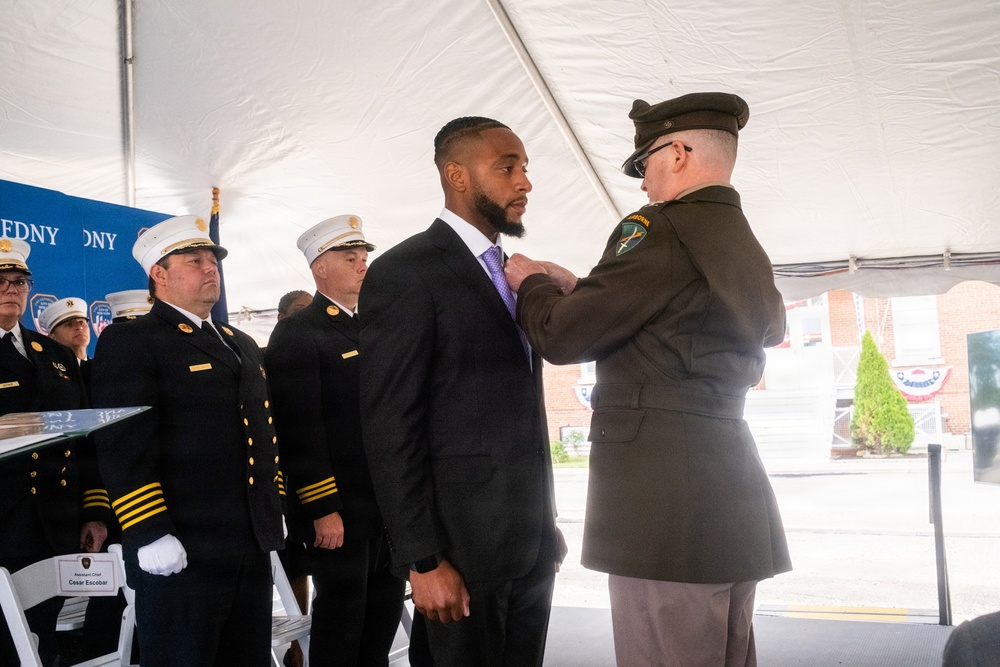 A formal awards ceremony is taking place under a white tent, where a military or law enforcement officer in olive/green dress uniform is pinning a medal or award onto a civilian man in a dark suit with a purple tie. Several FDNY (Fire Department of New York) officers in dark dress uniforms with white caps and gold insignia stand at attention in the background. A name placard reading 'César Escobar' is visible on a chair in the foreground.