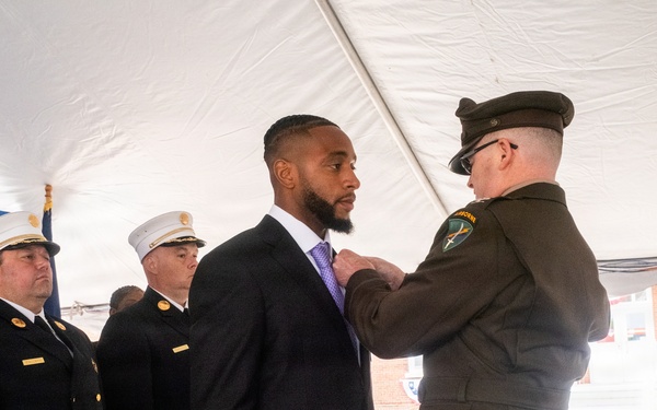 A formal awards ceremony is taking place under a white tent, where a military or law enforcement officer in olive/green dress uniform is pinning a medal or award onto a civilian man in a dark suit with a purple tie. Several FDNY (Fire Department of New York) officers in dark dress uniforms with white caps and gold insignia stand at attention in the background. A name placard reading 'César Escobar' is visible on a chair in the foreground.