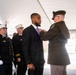 A formal awards ceremony is taking place under a white tent, where a military or law enforcement officer in olive/green dress uniform is pinning a medal or award onto a civilian man in a dark suit with a purple tie. Several FDNY (Fire Department of New York) officers in dark dress uniforms with white caps and gold insignia stand at attention in the background. A name placard reading 'César Escobar' is visible on a chair in the foreground.