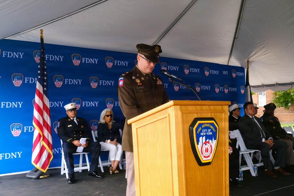 A military officer in U.S. Army dress uniform is speaking at a podium during an official FDNY (Fire Department City of New York) ceremony held under a white tent. The podium bears the FDNY shield emblem, and a repeating FDNY-branded backdrop is visible behind the stage. Several dignitaries and officials, including a uniformed FDNY officer, are seated on the stage behind the speaker.