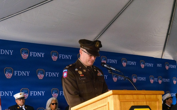 A military officer in U.S. Army dress uniform is speaking at a podium during an official FDNY (Fire Department City of New York) ceremony held under a white tent. The podium bears the FDNY shield emblem, and a repeating FDNY-branded backdrop is visible behind the stage. Several dignitaries and officials, including a uniformed FDNY officer, are seated on the stage behind the speaker.