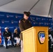 A military officer in U.S. Army dress uniform is speaking at a podium during an official FDNY (Fire Department City of New York) ceremony held under a white tent. The podium bears the FDNY shield emblem, and a repeating FDNY-branded backdrop is visible behind the stage. Several dignitaries and officials, including a uniformed FDNY officer, are seated on the stage behind the speaker.