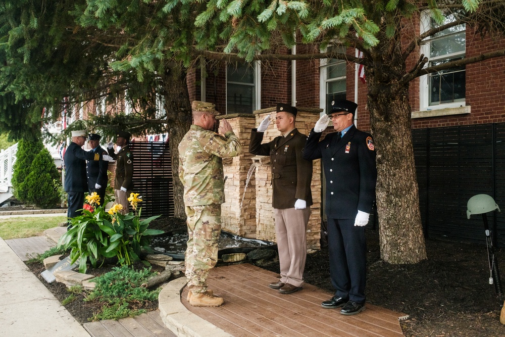 A military and first responder ceremony is taking place outdoors in front of a brick building. Multiple uniformed personnel are rendering salutes near what appears to be a memorial or monument with a small water feature. The gathering includes Army soldiers in various uniforms, a firefighter, and what appear to be Navy or other service members in the background.