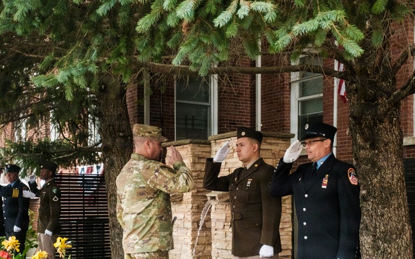 A military and first responder ceremony is taking place outdoors in front of a brick building. Multiple uniformed personnel are rendering salutes near what appears to be a memorial or monument with a small water feature. The gathering includes Army soldiers in various uniforms, a firefighter, and what appear to be Navy or other service members in the background.