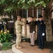 A military and first responder ceremony is taking place outdoors in front of a brick building. Multiple uniformed personnel are rendering salutes near what appears to be a memorial or monument with a small water feature. The gathering includes Army soldiers in various uniforms, a firefighter, and what appear to be Navy or other service members in the background.