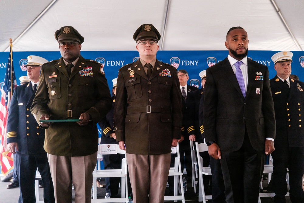 A formal FDNY ceremony is taking place under a white tent, featuring military personnel in U.S. Army dress uniforms alongside civilians and fire department officials. Three individuals stand prominently in the foreground: two Army officers in olive/brown dress uniforms with medals and insignia, and a civilian in a dark suit with a purple tie wearing a badge or pin. An American flag and FDNY-branded backdrop are visible in the background.