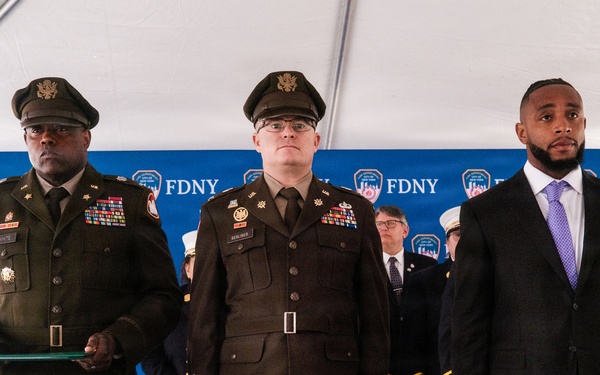 A formal FDNY ceremony is taking place under a white tent, featuring military personnel in U.S. Army dress uniforms alongside civilians and fire department officials. Three individuals stand prominently in the foreground: two Army officers in olive/brown dress uniforms with medals and insignia, and a civilian in a dark suit with a purple tie wearing a badge or pin. An American flag and FDNY-branded backdrop are visible in the background.