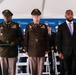 A formal FDNY ceremony is taking place under a white tent, featuring military personnel in U.S. Army dress uniforms alongside civilians and fire department officials. Three individuals stand prominently in the foreground: two Army officers in olive/brown dress uniforms with medals and insignia, and a civilian in a dark suit with a purple tie wearing a badge or pin. An American flag and FDNY-branded backdrop are visible in the background.