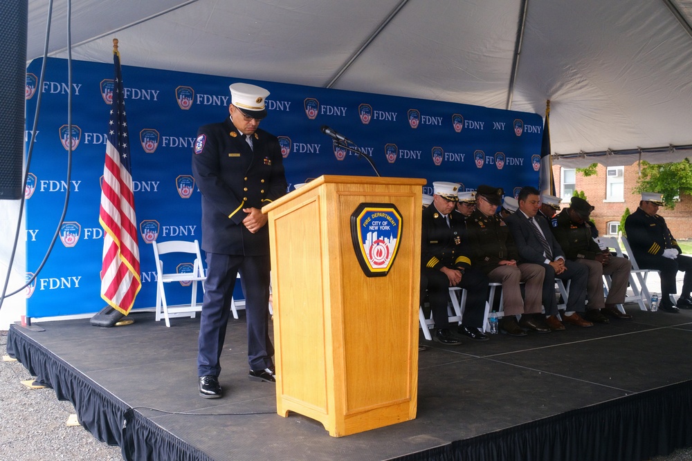 An FDNY (Fire Department City of New York) official in dress uniform stands at a podium during a formal ceremony, with his head bowed in what appears to be a moment of silence or prayer. Several other uniformed officials and dignitaries are seated on the stage behind him. The event is held under a tent with an FDNY-branded backdrop and an American flag visible on stage.