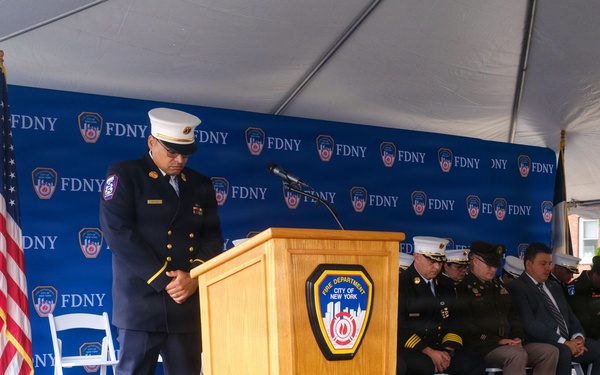 An FDNY (Fire Department City of New York) official in dress uniform stands at a podium during a formal ceremony, with his head bowed in what appears to be a moment of silence or prayer. Several other uniformed officials and dignitaries are seated on the stage behind him. The event is held under a tent with an FDNY-branded backdrop and an American flag visible on stage.