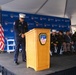 An FDNY (Fire Department City of New York) official in dress uniform stands at a podium during a formal ceremony, with his head bowed in what appears to be a moment of silence or prayer. Several other uniformed officials and dignitaries are seated on the stage behind him. The event is held under a tent with an FDNY-branded backdrop and an American flag visible on stage.