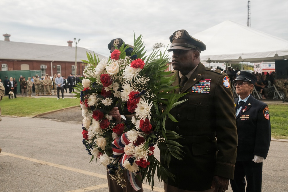 A military officer in dress uniform carries a large red, white, and blue floral wreath during what appears to be a formal memorial or commemorative ceremony. The officer displays numerous service ribbons and medals on his uniform, and is accompanied by other uniformed personnel including a firefighter. A crowd of attendees, including military personnel in camouflage uniforms, is visible in the background.