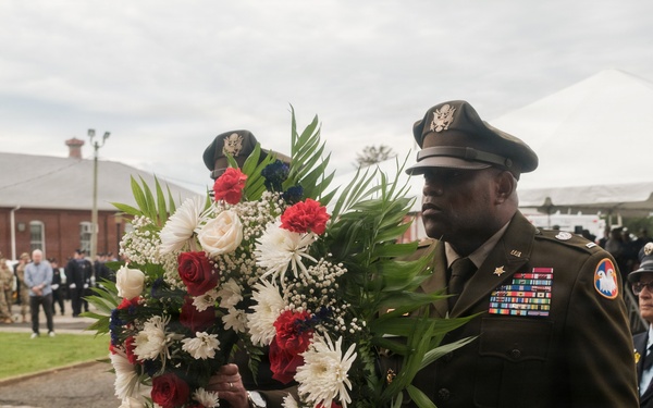 A military officer in dress uniform carries a large red, white, and blue floral wreath during what appears to be a formal memorial or commemorative ceremony. The officer displays numerous service ribbons and medals on his uniform, and is accompanied by other uniformed personnel including a firefighter. A crowd of attendees, including military personnel in camouflage uniforms, is visible in the background.