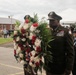 A military officer in dress uniform carries a large red, white, and blue floral wreath during what appears to be a formal memorial or commemorative ceremony. The officer displays numerous service ribbons and medals on his uniform, and is accompanied by other uniformed personnel including a firefighter. A crowd of attendees, including military personnel in camouflage uniforms, is visible in the background.