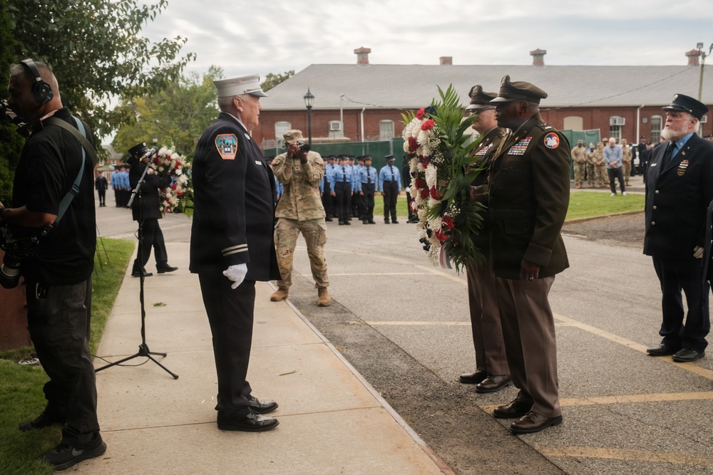 A formal memorial ceremony is taking place outdoors, likely a wreath-laying event. Military and fire department personnel in dress uniforms are participating, with two soldiers presenting a large floral wreath to a fire department officer. Rows of uniformed personnel are visible in the background, suggesting an organized ceremonial formation.