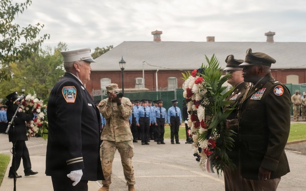 A formal memorial ceremony is taking place outdoors, likely a wreath-laying event. Military and fire department personnel in dress uniforms are participating, with two soldiers presenting a large floral wreath to a fire department officer. Rows of uniformed personnel are visible in the background, suggesting an organized ceremonial formation.