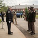 A formal memorial ceremony is taking place outdoors, likely a wreath-laying event. Military and fire department personnel in dress uniforms are participating, with two soldiers presenting a large floral wreath to a fire department officer. Rows of uniformed personnel are visible in the background, suggesting an organized ceremonial formation.