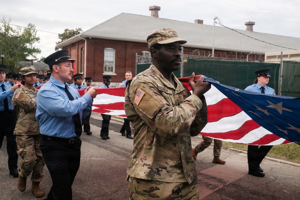 A group of U.S. Army soldiers in MultiCam/OCP camouflage uniforms and civilian first responders in blue uniforms are jointly carrying a large American flag during what appears to be a ceremonial event or parade. The participants are walking along a paved road near a red brick building surrounded by a security fence with razor wire. The scene suggests a formal military-civilian joint ceremony, possibly a memorial or patriotic observance.