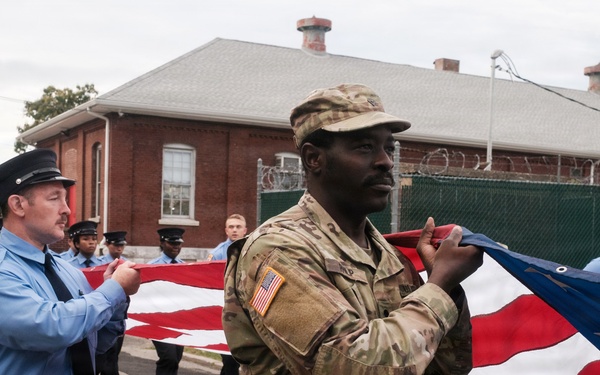 A group of U.S. Army soldiers in MultiCam/OCP camouflage uniforms and civilian first responders in blue uniforms are jointly carrying a large American flag during what appears to be a ceremonial event or parade. The participants are walking along a paved road near a red brick building surrounded by a security fence with razor wire. The scene suggests a formal military-civilian joint ceremony, possibly a memorial or patriotic observance.