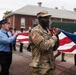A group of U.S. Army soldiers in MultiCam/OCP camouflage uniforms and civilian first responders in blue uniforms are jointly carrying a large American flag during what appears to be a ceremonial event or parade. The participants are walking along a paved road near a red brick building surrounded by a security fence with razor wire. The scene suggests a formal military-civilian joint ceremony, possibly a memorial or patriotic observance.