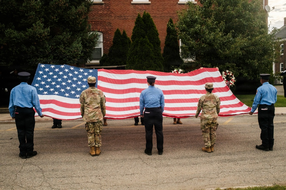 A group of military personnel and uniformed officers are holding a large American flag horizontally during what appears to be a ceremonial event. The scene takes place in a parking lot or open area in front of a brick building surrounded by trees. A floral arrangement is visible behind the flag, suggesting a memorial or commemorative ceremony.