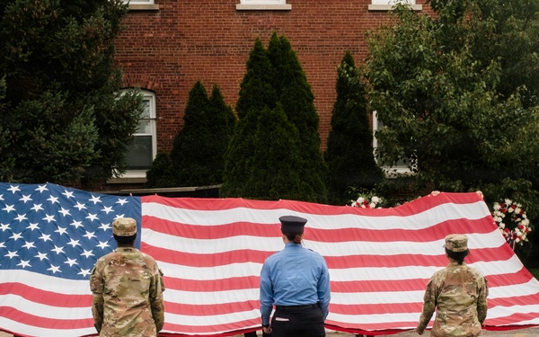 A group of military personnel and uniformed officers are holding a large American flag horizontally during what appears to be a ceremonial event. The scene takes place in a parking lot or open area in front of a brick building surrounded by trees. A floral arrangement is visible behind the flag, suggesting a memorial or commemorative ceremony.