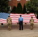 A group of military personnel and uniformed officers are holding a large American flag horizontally during what appears to be a ceremonial event. The scene takes place in a parking lot or open area in front of a brick building surrounded by trees. A floral arrangement is visible behind the flag, suggesting a memorial or commemorative ceremony.