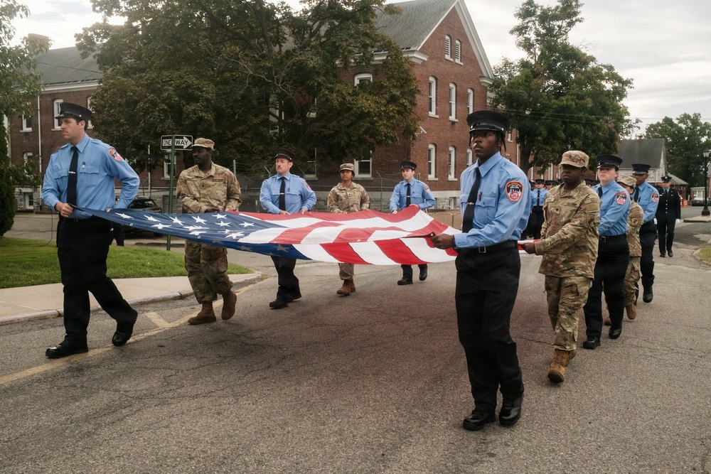 A group of uniformed personnel, including firefighters in light blue shirts and military soldiers in camouflage uniforms, are carrying a large American flag during what appears to be a ceremonial procession or parade. The event takes place on a street in front of a red brick building, suggesting a formal commemorative occasion. The mixed presence of fire department and military personnel indicates an inter-service ceremonial event.