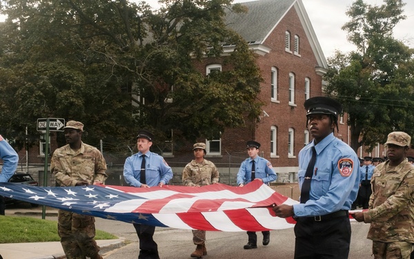 A group of uniformed personnel, including firefighters in light blue shirts and military soldiers in camouflage uniforms, are carrying a large American flag during what appears to be a ceremonial procession or parade. The event takes place on a street in front of a red brick building, suggesting a formal commemorative occasion. The mixed presence of fire department and military personnel indicates an inter-service ceremonial event.