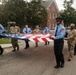 A group of uniformed personnel, including firefighters in light blue shirts and military soldiers in camouflage uniforms, are carrying a large American flag during what appears to be a ceremonial procession or parade. The event takes place on a street in front of a red brick building, suggesting a formal commemorative occasion. The mixed presence of fire department and military personnel indicates an inter-service ceremonial event.