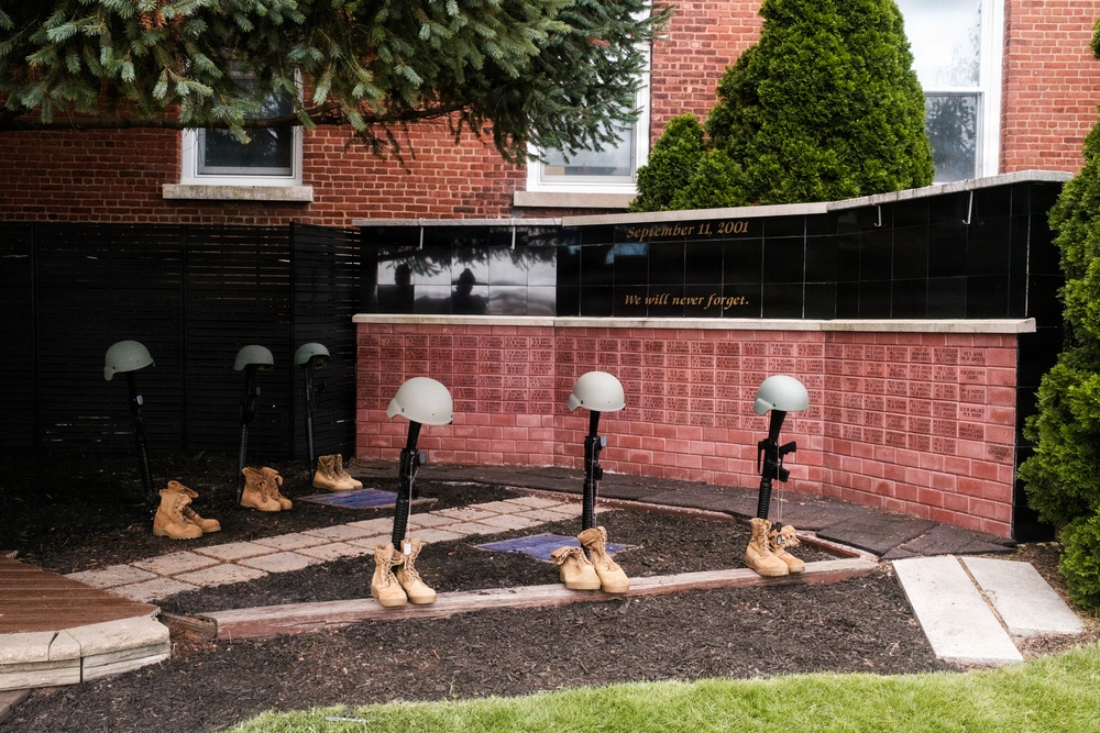 A memorial display commemorating September 11, 2001, featuring the 'Battlefield Cross' or 'Fallen Soldier Battle Cross' tribute — rifles with bayonets fixed into boots topped with helmets. The memorial is set against a brick wall engraved with names and inscribed with 'September 11, 2001' and 'We will never forget.' The display includes multiple such crosses arranged in a garden bed in front of what appears to be a public or civic building.