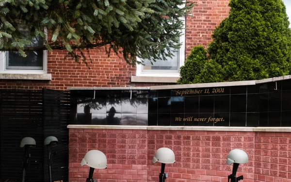 A memorial display commemorating September 11, 2001, featuring the 'Battlefield Cross' or 'Fallen Soldier Battle Cross' tribute — rifles with bayonets fixed into boots topped with helmets. The memorial is set against a brick wall engraved with names and inscribed with 'September 11, 2001' and 'We will never forget.' The display includes multiple such crosses arranged in a garden bed in front of what appears to be a public or civic building.